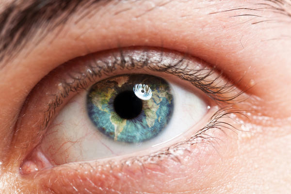 Closeup of a blue eye with hues of yellow and green mixed in, reflection of light on the near the top of the pupil.